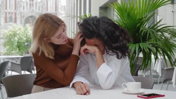 Jonge vrouw troost haar huilende vriend terwijl ze in een café op straat zit. Vrouwelijke vriendschap. Het ene meisje huilt, het andere troost haar. — Stockvideo