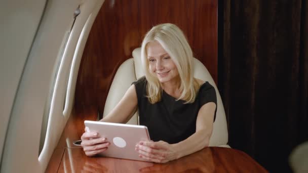 Смиллинг зрелой бизнесвумен с помощью планшетного компьютера во время путешествия на частном реактивном самолете. Богатая пожилая женщина смотрит контент на гаджете и наслаждается роскошными путешествиями на авиакомпании первого бизнес-класса — стоковое видео