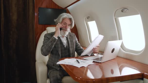 Homme d'affaires mature barbu occupé travaillant avec un ordinateur portable lisant des documents papier et parlant sur un téléphone portable tout en voyageant en jet privé ou en avion de première classe d'affaires — Video