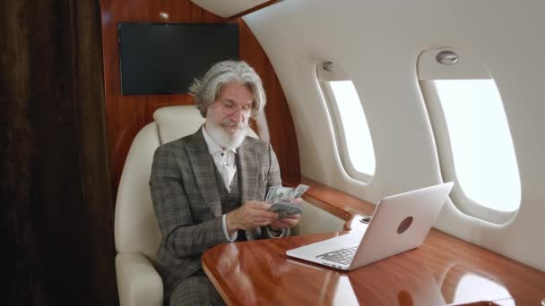 Heureux homme riche senior compte de l'argent tout en volant dans un jet privé. Un millionnaire masculin souriant qui réussit se réjouit de ses profits, raconte en argent comptant, lors d'un vol de première classe — Video