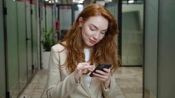 Close-up glimlachende aantrekkelijke zakenvrouw met behulp van smartphone. Portret van een zelfverzekerde vrouw executive met rood haar in formele slijtage met behulp van app op mobiele telefoon tijdens het staan in kantoor gang. — Stockvideo