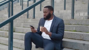 Sakallı Afrikalı Amerikalı adam modern bir akıllı telefonla internette geziniyor ve ofis merkezinin yanındaki merdivenlerde otururken kahve içiyor..
