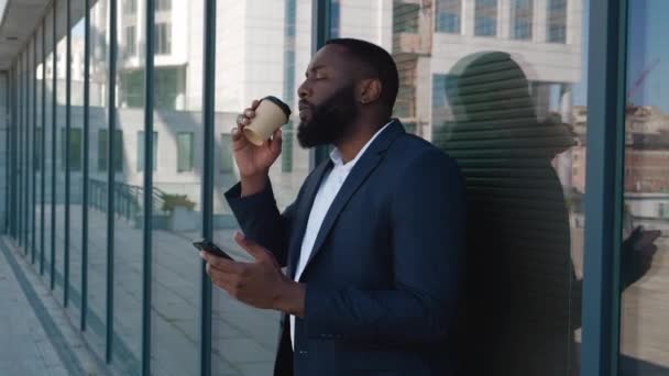 Portret afrykańskiego biznesmena stojącego z kawą i telefonem na ulicy. mężczyzna biurowy pracownik jest picie kawa w przerwie i przeglądanie jego smartfon w pobliżu nowoczesny duży biurowiec — Wideo stockowe