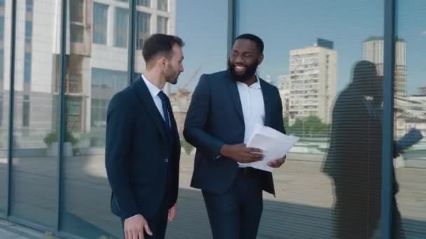 Resmi takım elbiseli iki güler yüzlü iş adamı ellerinde kağıt parçalarıyla şehir merkezindeki ofis binasının yakınındaki iş yerlerinde yürüyor ve tartışıyorlar. Girişimci erkekler açık havada konuşuyor. — Stok video