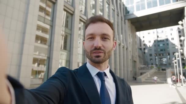 Sonriente joven hombre de negocios haciendo videollamada selfie de pie al aire libre en el distrito de negocios. Retrato de un bloguero de negocios filmando video blog al aire libre cerca del moderno edificio de oficinas. — Vídeos de Stock
