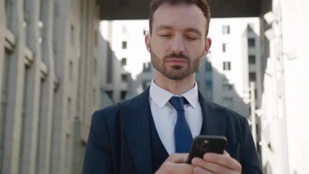 Portrét atraktivního podnikatele pomocí chytrého telefonu stojící v centru u Moderní kancelářské budovy. Nosí klasický oblek. Sociální síť. Aplikace. Smartphone. — Stock video