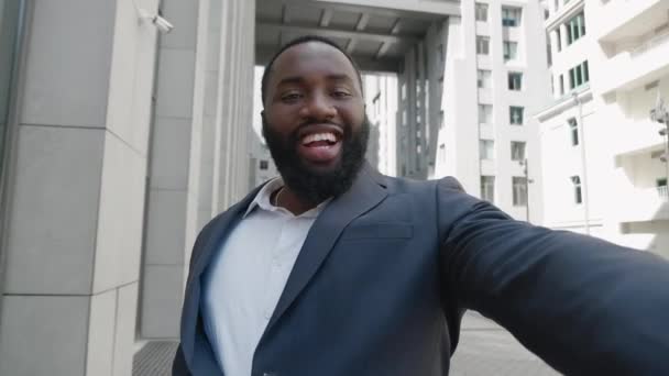 アフリカ系アメリカ人のビジネスマンを笑顔にして、ビジネス街の屋外に立ってビデオ通話をします。現代のオフィスビルの近くに屋外でビデオブログを撮影ビジネスマンブロガーの肖像. — ストック動画