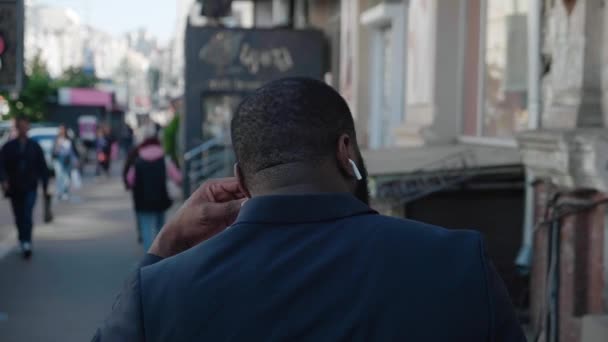 Vista trasera trasera del hombre afroamericano con auriculares inalámbricos caminando por la calle en la gran ciudad en cámara lenta — Vídeo de stock