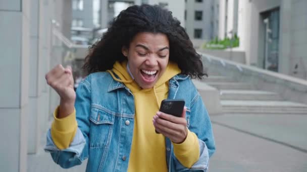 Mujer afroamericana feliz mirando en el teléfono inteligente se sienten sorprendidos, diciendo WOW y hace sí gesto. Celebrando la victoria del éxito. Emocionado ganador negro femenino mantener el teléfono móvil sorprendido con buenas noticias. — Vídeo de stock