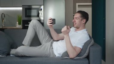 Akıllı telefonlu gülümseyen genç adam oturma odasında kanepede uzanırken video görüşmesi yapıyor. Evdeki kanepede dinlenen mutlu bir adamın portresi.