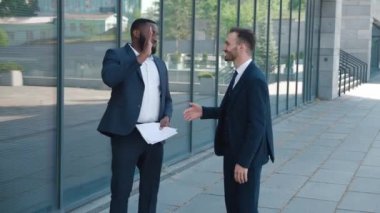 Resmi takım elbiseli, gülümseyen iki iş adamı iş konuşuyorlar, zaferden ve beşlik çakmaktan memnunlar, şehir merkezinde ofis binasının yanında duruyorlar. Erkek girişimciler dışarıda konuşuyor..