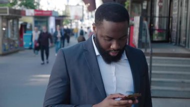 Kendine güvenen sakallı Afrikalı adam şehir sokağında beklerken modern akıllı telefondan internete göz atıyor. Dışarıda cep telefonu kullanarak resmi takım elbise giyen iş adamının portresi. Şehir manzarası.