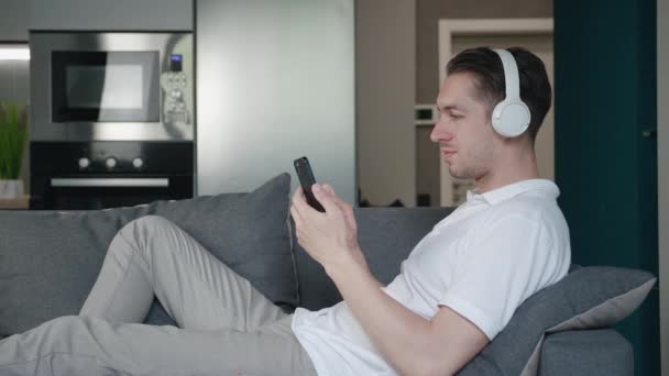 Portrét veselého mladého muže ležícího na pohovce a používajícího aplikaci smartphonu, který poslouchá hudbu s bílými sluchátky, zatímco odpočívá doma na gauči. — Stock video