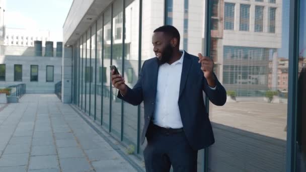 Aufgeregt schaut ein afrikanisch-amerikanischer Geschäftsmann auf sein Smartphone und unterschreibt. Glücklicher Mann Unternehmer im Anzug tanzt Siegertanz feiert Erfolg Sieg oder erfolgreichen Deal — Stockvideo