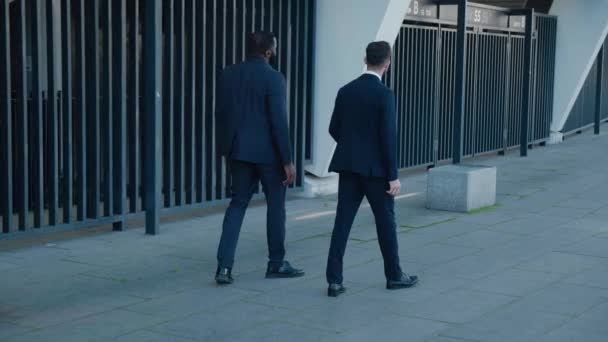 Vista posteriore sui due uomini d'affari multietnici in abiti formali passeggiando per la strada nella moderna città degli affari e parlando. Posteriore. — Video Stock
