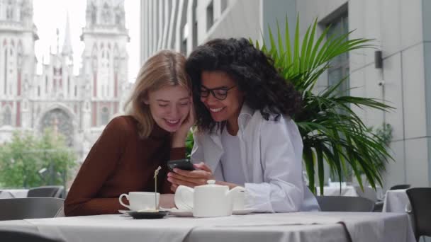 Zwei junge schöne Freundinnen sitzen draußen im Café, schauen aufs Telefon und unterhalten sich, lachen. Zwei unterschiedlich schöne Weibchen nutzen Social Media auf dem Smartphone im Café, moderne Generation — Stockvideo