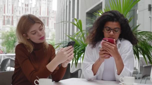 Two bored diverse women friends browse the internet on their smartphones at outdoors cafe. — Stock Video