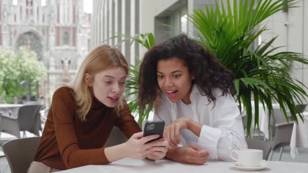 Dos amigas disfrutan del éxito en el teléfono móvil en la cafetería de la calle. Chicas sorprendidas celebrando la victoria en el teléfono inteligente, disfrutando de ganar premios, mientras se sientan en la cafetería al aire libre — Vídeo de stock