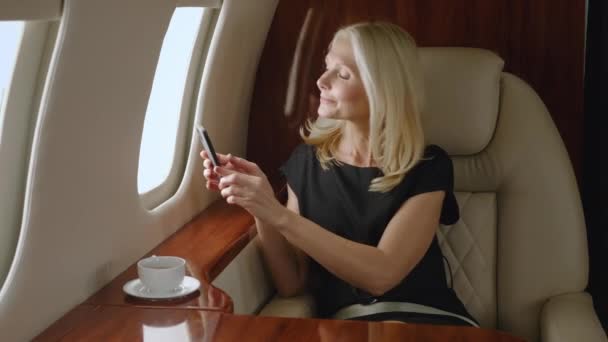 Belle femme mûre millionnaire prenant des photos avec smartphone depuis la fenêtre de l'avion. Femme d'affaires voyageant en jet privé ou en première classe affaires. Voyage de luxe. — Video