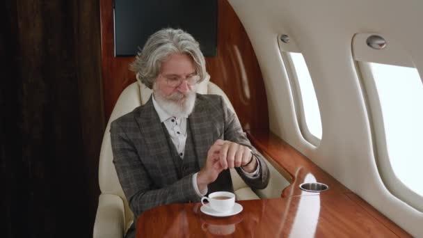 Busy mature business man looking on his watch and then at window while travels on private plane. Rich senior man drinking tea or coffee while enjoying luxury journey in first business class airlines — Stock Video