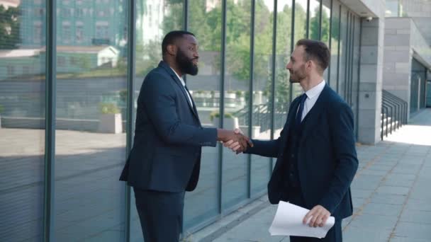 2人のビジネスマンが会うとき握手をしたり、取引に同意したり、挨拶をしたりします。取引を行う2人の笑顔のビジネスマンの握手の肖像画。背景にオフィスビル。ビジネス街. — ストック動画