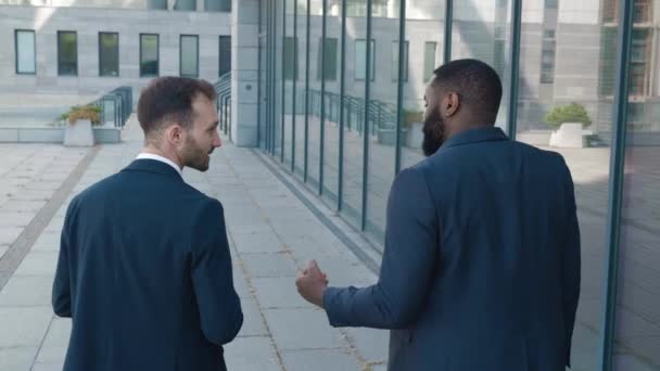 Retrovisore posteriore di due colleghi uomini sorridenti in abiti formali chattare sulla strada per il lavoro. Uomini d'affari africani e caucasici che parlano mentre camminano in centro. — Video Stock