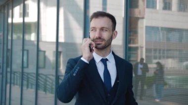 Resmi takım elbiseli, kendine güvenen sakallı iş adamı iş merkezinde yürürken telefonla konuşuyor..