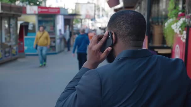 Πίσω όψη του Αφροαμερικανού επιτυχημένου άνδρα με κοστούμι που περπατάει στο δρόμο της πόλης και μιλάει από κινητό τηλέφωνο. Αστικό τοπίο. Οπίσθιος επιχειρηματίας με αυτοπεποίθηση απαντά στο τηλέφωνο έξω, ενώ μεταβαίνει στην εργασία — Αρχείο Βίντεο