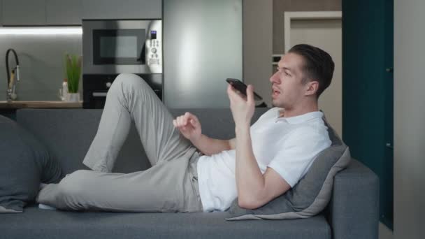 スピーカーフォンを使ってスマホを持っているソファに腰を下ろしている若者。自宅のリビングルームで居心地の良いソファに横たわっている間に音声メッセージを記録するために携帯電話を使用して陽気な男性. — ストック動画