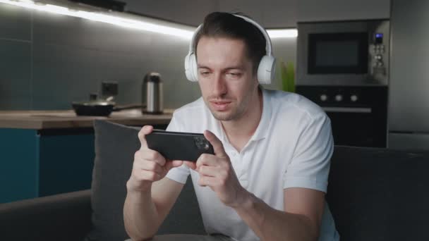 Közelkép a pihentető fiatalember fülhallgató ül a kanapén néz videó okostelefon a modern lakás — Stock videók