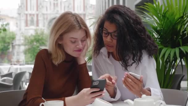 Two young beautiful women friends sitting in cafe outdoors, looking at phones and talking, laughing. Two diverse beautiful females use social media on smartphones in coffee shop, modern generation — Stock Video