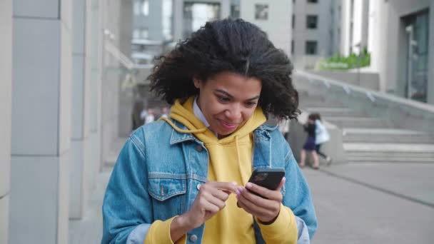 Hermosa joven afroamericana mujer de pie al aire libre y mensajes de texto en el teléfono móvil y sonriendo. Sonriente estudiante en chaqueta de jean tocando y desplazándose en el teléfono inteligente en la ciudad. — Vídeo de stock