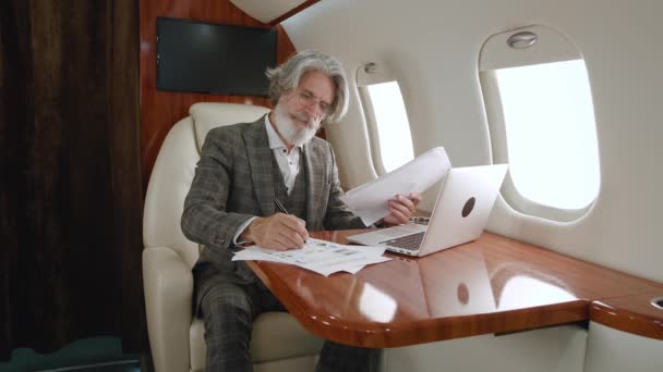 Homme mûr barbu millionnaire travaillant sur un ordinateur portable lisant des documents papier tout en voyageant en avion. Portrait d'un homme d'affaires senior en jet privé ou en première classe d'affaires avec ordinateur — Video