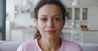 4K video, beyaz ahşap mutfak arka planındaki koltukta oturan orta doğulu sakin bir kadının kamera görüntüsü. Olgun, melez bir kadının yakın plan portresi. 4K video görüntüleri.