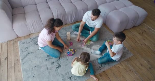 Pais com crianças focadas na construção de forma atmosférica a partir de construtor magnético colorido, sentado no chão da sala de estar. Imagens de vídeo, 4 K. — Vídeo de Stock