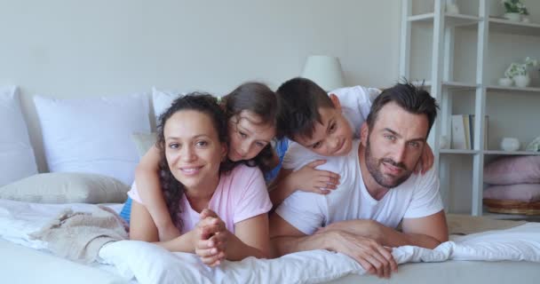 Multirazziale famiglia felice sdraiata sul letto e sorriso carino alla macchina fotografica. Ritratto di famiglia di bambini felici adorabili che abbracciano i loro genitori. Amore, affetto e concetti di infanzia spensierata. Video 4K — Video Stock