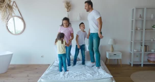 Βίντεο 4K με χαρούμενους μεσήλικες γονείς να πηδάνε στο κρεβάτι με τα αξιολάτρευτα παιδιά τους, το σχολιαρόπαιδο και το μικρότερο κορίτσι. Full length ξέγνοιαστη οικογένεια δύο γενιές — Αρχείο Βίντεο