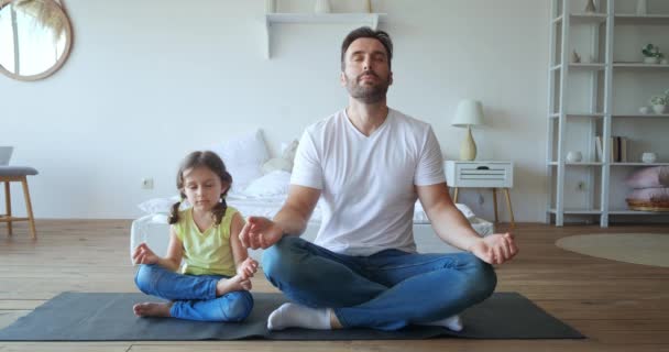 Menina bonito meditando com seu pai bonito, sentado em posição de lótus no tapete de ioga no quarto. Imagens de vídeo 4K. Educação, estilo de vida saudável, parentalidade e conceitos de infância. — Vídeo de Stock