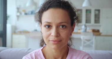 4K video görüntüleri. Çekici Afrikalı Amerikalı kadın beyaz ahşap mutfak arka planındaki koltukta oturan kameraya bakıyor. Olgun bir melez kadının yakın plan portresi.