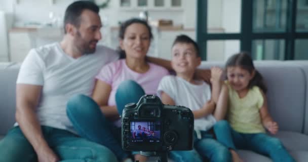 Οικογένεια στον καναπέ και καταγραφή βίντεο blog στην κάμερα τοποθετείται σε τρίποδο με θολή φόντο στο σαλόνι. Κίνηση, δραστήρια χειρονομίες, χαμόγελα, γέλια και διασκέδαση. — Αρχείο Βίντεο