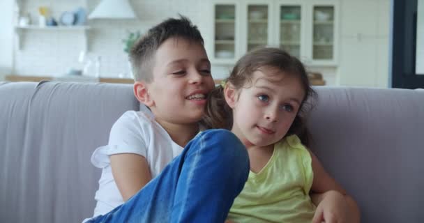 En omsorgsfuld bror krammer sin lillesøster, taler med hende, sidder på sofaen i stuen. 4K video af glade familieforhold med venskab, kærlighed og omsorg – Stock-video