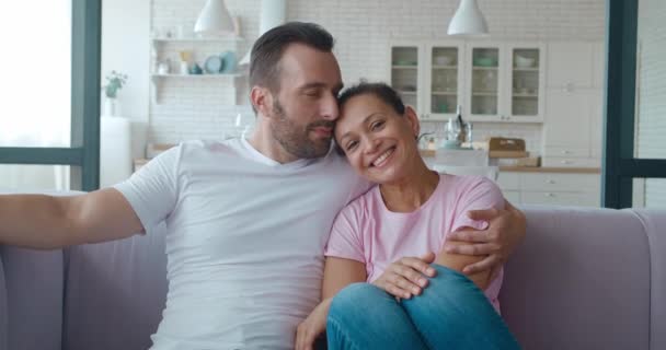 Bonito homem carinhosamente abraçando e beijando sua esposa, gostando de passar tempo juntos no sofá na sala de estar. Filmagem de vídeo 4K sobre o feliz casal multirracial de meia idade — Vídeo de Stock