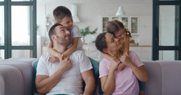 Bambini adorabili abbracciano i loro genitori amorevoli seduti sul divano, poi lo scolaro abbraccia teneramente la sua famiglia. Video in 4K su felici relazioni familiari. Amore, tenerezza e affetto concetto. — Video Stock