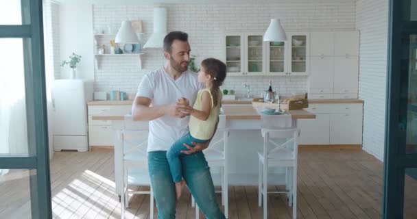 Video de un padre sonriente y su hija pequeña disfrutando de pasar el fin de semana juntos. Linda niña con dos colas de caballo sentada en los brazos del padre y bailando en la cocina. — Vídeos de Stock