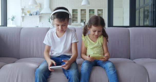 Bother and sister enjoy multimedia games. Cute little girl smiles, looking at mobile phone, charming boy in headphones actively plays computer games sitting on sofa in living room. 4 K video — 비디오