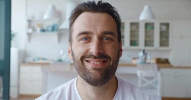 4K video. Yakışıklı, olgun, sakallı, dişlek gülüşüyle gülümseyen ve kamera karşısında mutfak arka planına bakan Avrupalı bir adamın portresi. Yakışıklı beyaz bir adamın portresi.