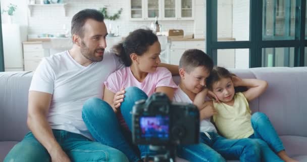 Los bloggers familiares multirraciales saludan mirando la cámara montada en un trípode, gesticulando activamente, sonriendo, comunicándose y divirtiéndose durante la grabación del video blog — Vídeo de stock