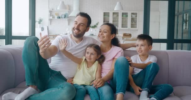 Beau homme, père, assis à côté de sa femme et de ses enfants, tenant un téléphone portable et parlant par appel vidéo. Famille saluant bonjour en regardant l'écran du téléphone portable — Video