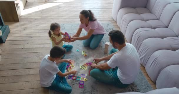 Família feliz aproveitando o tempo com as crianças jogando construtor magnético sentado no chão do sofá. Um pai de meia-idade ajuda seus filhos a construir formas geométricas. Imagens de vídeo, 4K — Vídeo de Stock