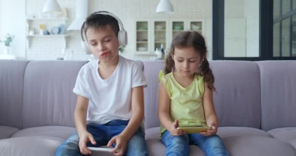 4 K videofelvétel imádnivaló iskolás fiúról fejhallgatóval aktív zenét hallgat és táncol a kanapén ülve, csinos, fiatalabb lány két lófarokkal és mobilon nézi. — Stock videók